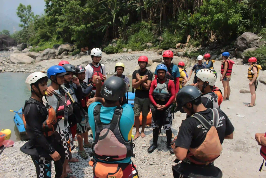 A group of kayakers wearing helmets and life jackets gathers for a safety briefing before kayaking.